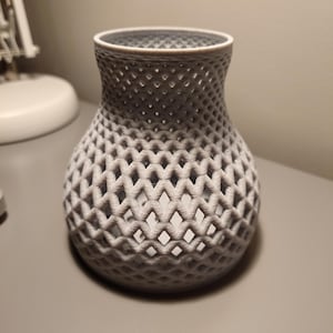 May include: A grey, 3D printed vase with a geometric, lattice design. The vase has a round base and a wide opening.