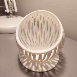 May include: A white, 3D printed, egg-shaped object with a lattice design. The object has a round base and is sitting on a gray surface.