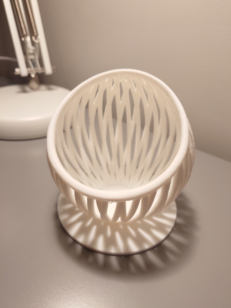May include: A white, 3D printed, egg-shaped object with a lattice design. The object has a round base and is sitting on a gray surface.