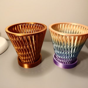 Può includere: Due vasi decorativi stampati in 3D, uno di colore rame e l'altro con un gradiente di blu, verde e arancione.