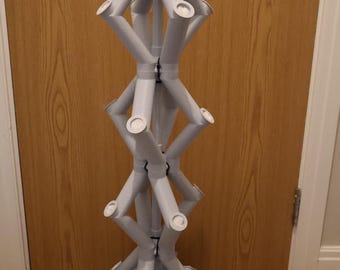 Peut inclure: Une lampe géométrique à plusieurs niveaux, de couleur blanche, imprimée en 3D, avec une base carrée et un cordon. La lampe est composée d'une série de formes cylindriques reliées entre elles qui forment une tour.