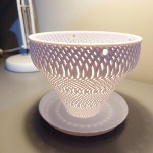 Op de afbeelding: Witte keramische plantenpot met een geometrisch tralieontwerp. De pot heeft een brede, ondiepe basis en een hoge, conische bovenkant. Het tralieontwerp laat licht door de pot schijnen.