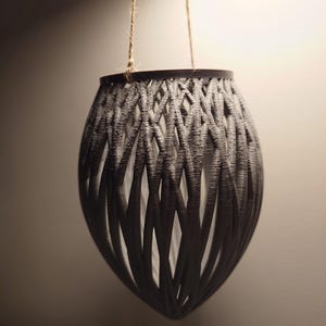May include: A grey, woven hanging planter with a rope loop for hanging. The planter is made of a woven material that resembles a basket.