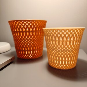 May include: Two orange plastic plant pots with a lattice design. The larger pot is a darker orange and the smaller pot is a lighter orange.