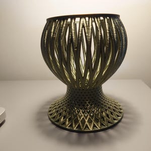 May include: A gold-coloured metal vase with a geometric design. The vase has a wide, round base and a narrow, cylindrical neck.