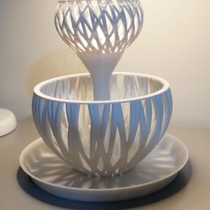 Könnte beinhalten: Eine weiße dekorative Lampe mit einem einzigartigen Design. Die Lampe hat einen schalenförmigen Sockel mit einem Gittermuster, einen zentralen Stiel und eine Oberseite mit einem ähnlichen Gitterdesign. Die Lampe steht auf einer runden, weißen Platte.