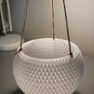 Könnte beinhalten: Ein weißer, runder, hängender Pflanztopf mit einer strukturierten Oberfläche. Der Topf ist aus einem Material, das wie Plastik oder Keramik aussieht. Er hängt an einer braunen Schnur.