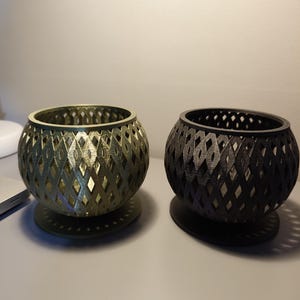 May include: Two spherical candle holders with a diamond lattice design. One is gold, the other black. They sit on circular bases. The candle holders are made of a 3D printed material. The diameter of each is approximately 12 cm.