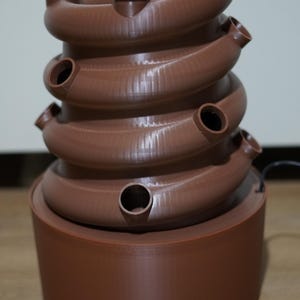 Könnte beinhalten: Ein brauner, selbstbewässernder Pflanzentopf aus Kunststoff mit mehreren Etagen und einem runden Sockel. Der Topf hat mehrere kleine Löcher zum Pflanzen.