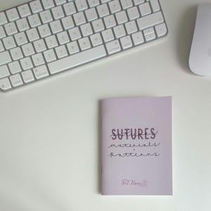 May include: A light purple booklet with the text SUTURES materials + patterns and a small logo at the bottom, positioned near a white computer keyboard and a white computer mouse. The cover features dark text in a handwritten style font.
