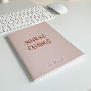 May include: A light pink notebook with the text NURSE CLINICS printed in bold brown letters with a thin black cursive line overlay. A small logo with the text Vet Nurse Life and a line drawing of a dog is visible at the bottom of the cover.