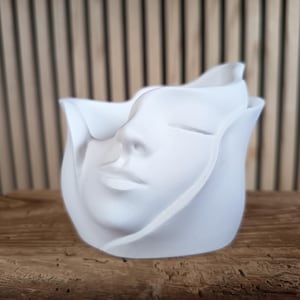Könnte beinhalten: Ein weißer Keramik-Pflanztopf in Form eines stilisierten Gesichts mit geschlossenen Augen und zarten Gesichtszügen. Der Pflanztopf hat ein blütenblattartiges Design um das Gesicht. Er steht auf einer Holzoberfläche.
