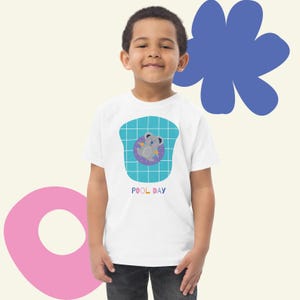 Puede incluir: Una camiseta blanca con un diseño de koala de dibujos animados en una boya de piscina. El koala es gris con una boya morada, sobre un fondo de cuadrícula azul. Las palabras "POOL DAY" están impresas debajo.