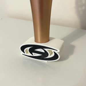 May include: A white and black decorative holder with a bronze-colored object inserted. The holder features a stylized black design on the front. The bronze object is cylindrical.