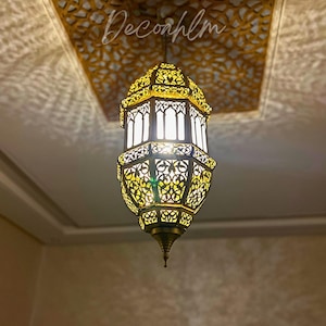 May include: Ornate, gold-colored pendant light with intricate cut-out patterns. The light fixture hangs from a ceiling with a matching decorative design. The light is illuminated, casting shadows. The text "Decorhlm" is visible on the ceiling.