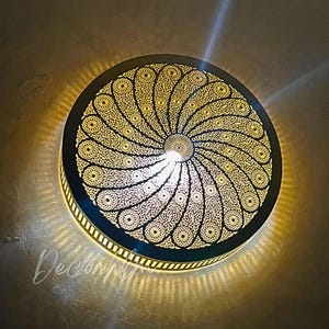 May include: A round, ornate, gold-colored metal wall sconce with a detailed, openwork design. The intricate pattern radiates from the center, creating a sunburst effect. The sconce is illuminated, casting a warm glow and intricate shadows on the wall.