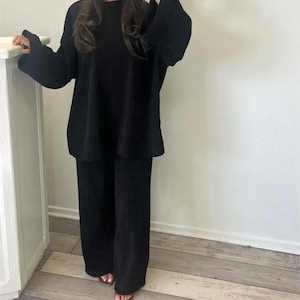 May include: A black two-piece outfit with a long-sleeved top and wide-leg pants. The top has a relaxed fit and a round neckline. The pants have a pleated design.