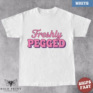 May include: White t-shirt with the text "Freshly Pegged" in pink and purple lettering. The shirt has short sleeves and a crew neck. The Bold Print Clothing Store logo is at the bottom left.