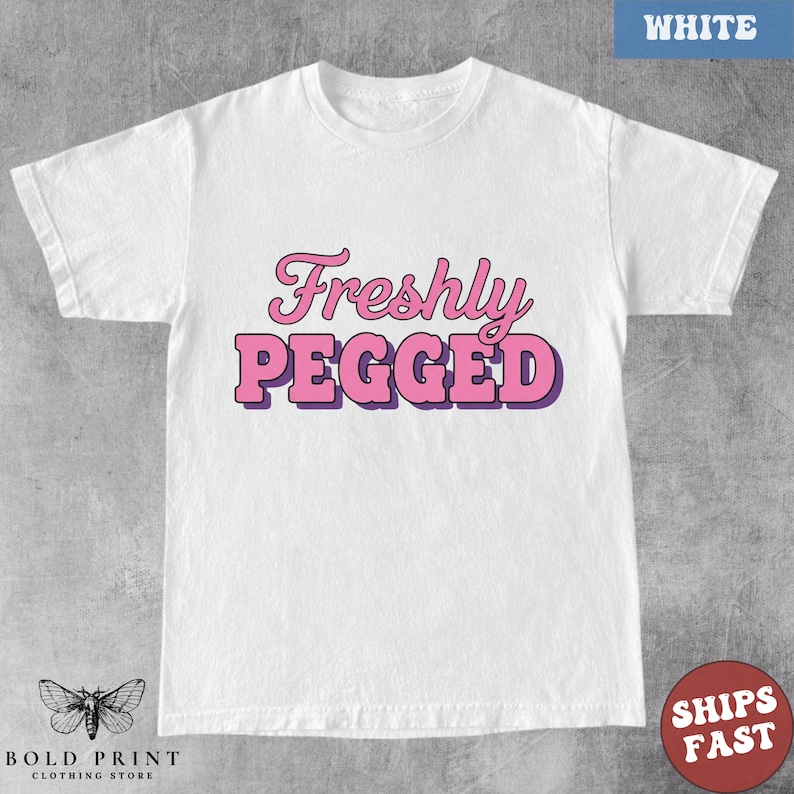 May include: White t-shirt with the text "Freshly Pegged" in pink and purple lettering. The shirt has short sleeves and a crew neck. The Bold Print Clothing Store logo is at the bottom left.
