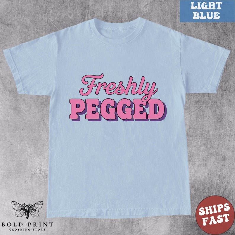 May include: Light blue t-shirt with the text "Freshly Pegged" in pink, outlined in purple. The shirt has short sleeves and a crew neck. The brand logo is at the bottom left corner, and a "Ships Fast" sticker is on the bottom right.