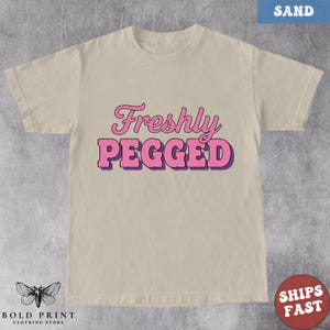 May include: Sand-colored t-shirt with the text "Freshly Pegged" in pink and purple lettering. The shirt has a crew neck and short sleeves. The Bold Print Clothing Store logo is at the bottom left.