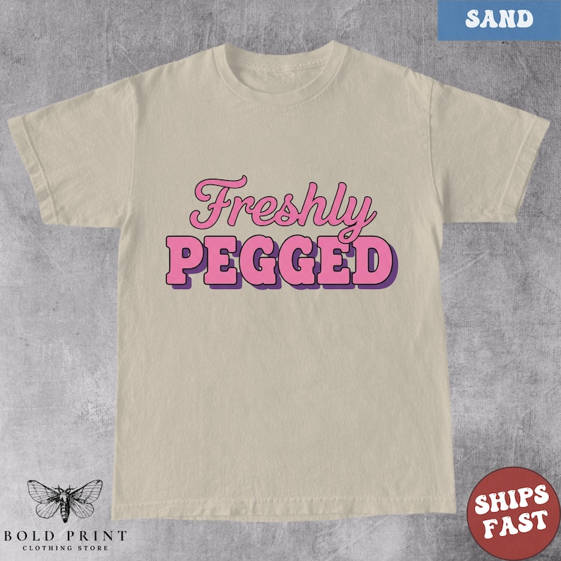 May include: Sand-colored t-shirt with the text "Freshly Pegged" in pink and purple lettering. The shirt has a crew neck and short sleeves. The Bold Print Clothing Store logo is at the bottom left.