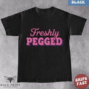 May include: Black t-shirt with the text "Freshly Pegged" in pink letters with a purple outline. The shirt has a crew neck and short sleeves. The words "Bold Print Clothing Store" and a butterfly logo are at the bottom.