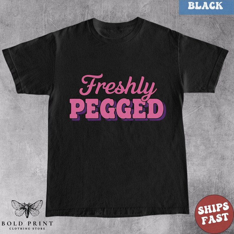 May include: Black t-shirt with the text "Freshly Pegged" in pink letters with a purple outline. The shirt has a crew neck and short sleeves. The words "Bold Print Clothing Store" and a butterfly logo are at the bottom.