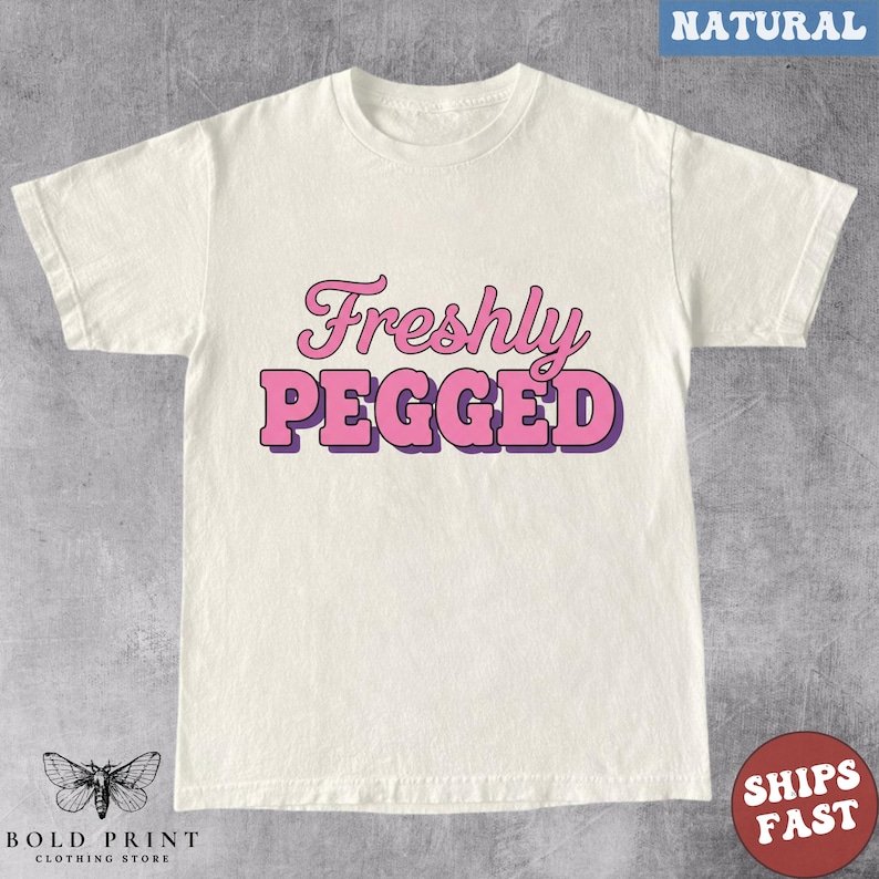 May include: Cream-colored t-shirt with the text "Freshly Pegged" in pink and purple lettering. The shirt has a crew neck and short sleeves. The Bold Print Clothing Store logo is at the bottom.