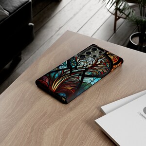May include: A black phone case with a colorful stained glass design featuring a tree with intricate branches and leaves. The design is in shades of blue, green, red, yellow, and orange.