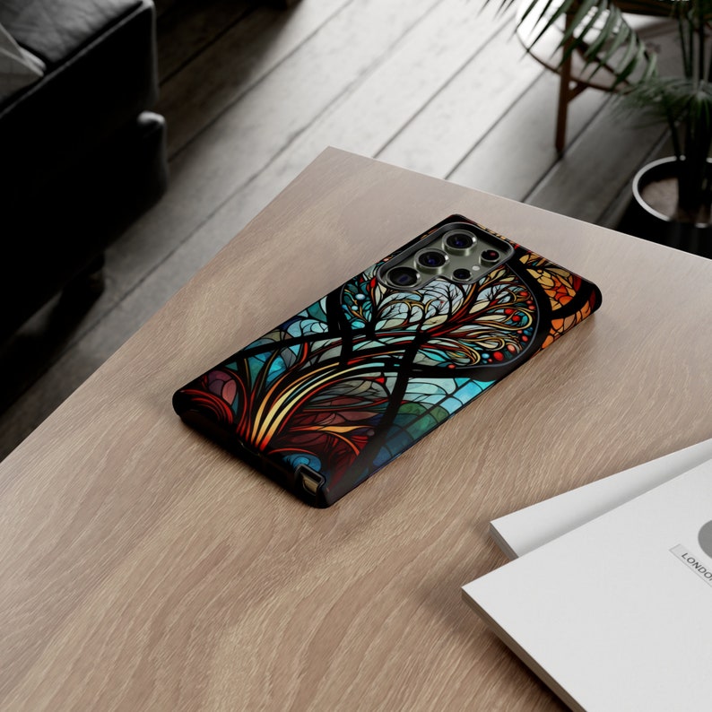 May include: A black phone case with a colorful stained glass design featuring a tree with intricate branches and leaves. The design is in shades of blue, green, red, yellow, and orange.