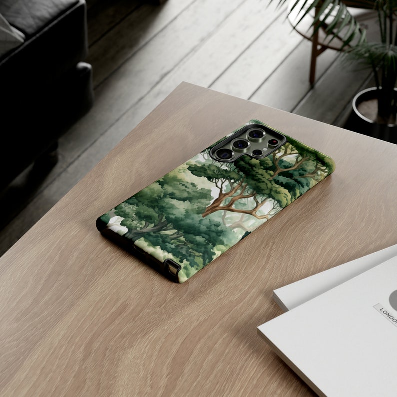 Peut inclure: Coque de smartphone avec une peinture &agrave; l'aquarelle d'une for&ecirc;t, avec des arbres et des branches verts. Le bo&icirc;tier a un bord noir et une ouverture pour l'appareil photo. Le design &eacute;voque la nature.
