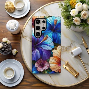 May include: A smartphone case featuring a vibrant floral design with blue, purple, and orange hibiscus flowers. The case has a glossy finish and is designed to fit a specific phone model. The flowers are arranged in a stained-glass style.