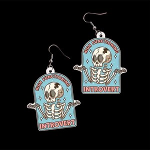 Pu&ograve; includere: Un paio di orecchini pendenti con un design a scheletro di cartone animato. Ogni orecchino ha uno sfondo azzurro con le parole "HIGH FUNCTIONING INTROVERT" e un'illustrazione di uno scheletro. Gli orecchini hanno ganci color argento.