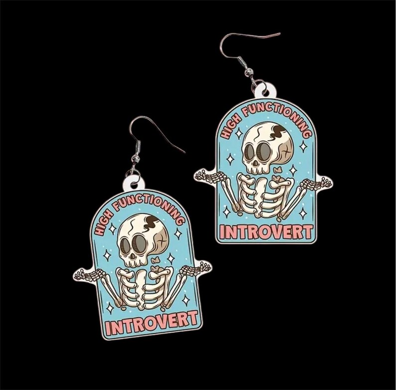 Pu&ograve; includere: Un paio di orecchini pendenti con un design a scheletro di cartone animato. Ogni orecchino ha uno sfondo azzurro con le parole "HIGH FUNCTIONING INTROVERT" e un'illustrazione di uno scheletro. Gli orecchini hanno ganci color argento.