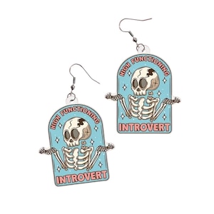 May include: A pair of novelty earrings featuring a cartoon skeleton design. Each earring has a light blue background with the words "HIGH FUNCTIONING" and "INTROVERT" in pink. The skeleton is white with black details.