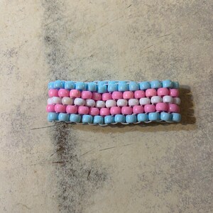 Puede incluir: Una pulsera de cuentas con un patrón de cuentas celeste, rosa y blancas. Las cuentas están dispuestas en filas horizontales, creando un efecto rayado. La pulsera está hecha de pequeñas cuentas redondas ensartadas juntas.