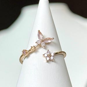 Könnte beinhalten: Goldring mit Schmetterlings- und Blumenmotiv. Der Schmetterling hat rosa und weiße Steine, die Blume hat klare Steine.