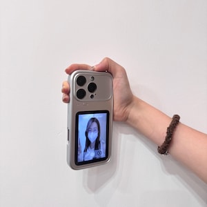 May include: Silver phone case with a camera-style design, featuring multiple lenses and a small screen displaying a person wearing a face mask. The case is held in a hand against a white background.