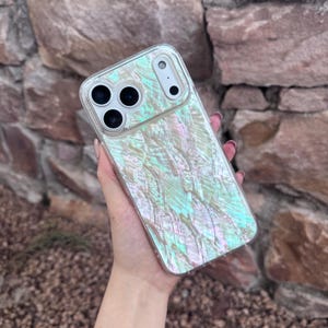 以下が含まれることがあります： 透明な携帯電話ケース。イリデッセントで真珠のようなデザイン。ケースは、緑、ピンク、青の色合いが混ざった、質感のある輝く表面を持っています。ケースには、電話のカメラとボタン用の切り欠きがあります。