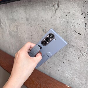 May include: A light blue phone case with a built-in stand and a loop grip. The case has a camera module with three lenses. The grip has a repeating circular design. The phone case is held against a gray concrete wall.