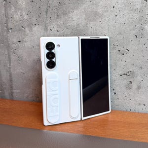 May include: White phone case for a foldable phone. The case has a camera module with three lenses and the word "FOLD" embossed on the side. The phone screen is black. The case is on a wooden surface.