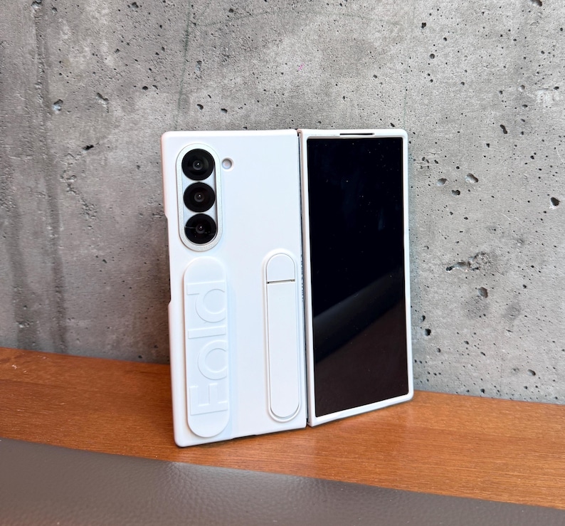 May include: White phone case for a foldable phone. The case has a camera module with three lenses and the word "FOLD" embossed on the side. The phone screen is black. The case is on a wooden surface.