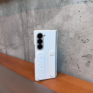 May include: A white phone case with a triple-lens camera and the word "Fold" embossed on the side. The case has a built-in stand and is designed to protect a foldable phone. The case is on a wooden surface.