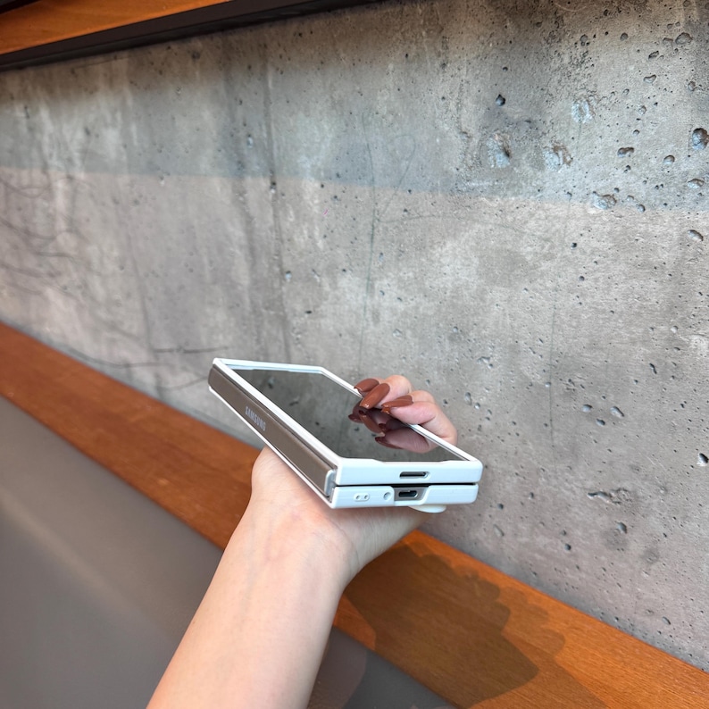 May include: A white and brown smartphone case with a reflective screen. The case has a rectangular shape with rounded corners. The phone case is held in a hand.