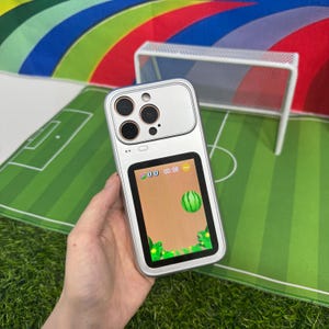 May include: Silver phone case with a built-in screen displaying a retro video game with a watermelon. The case has a rectangular screen area and a camera module with multiple lenses. The background features a soccer field.