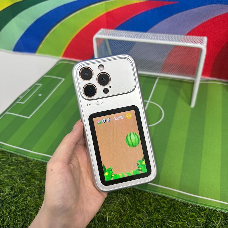 May include: Silver phone case with a built-in screen displaying a retro video game with a watermelon. The case has a rectangular screen area and a camera module with multiple lenses. The background features a soccer field.