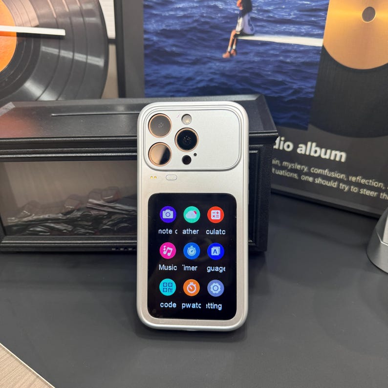 May include: Silver smartphone with multiple camera lenses and a small screen displaying app icons. The screen shows the words "note", "cather", "culato", "Music", "Timer", "guage", "code", "pwatc", and "itting".