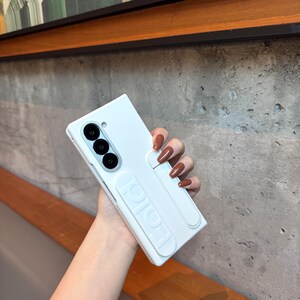 May include: A light blue phone case with a built-in stand. The case has three camera lenses and a small circular lens. The stand is white and has the word "HOLD" embossed on it. The case is held by a person.