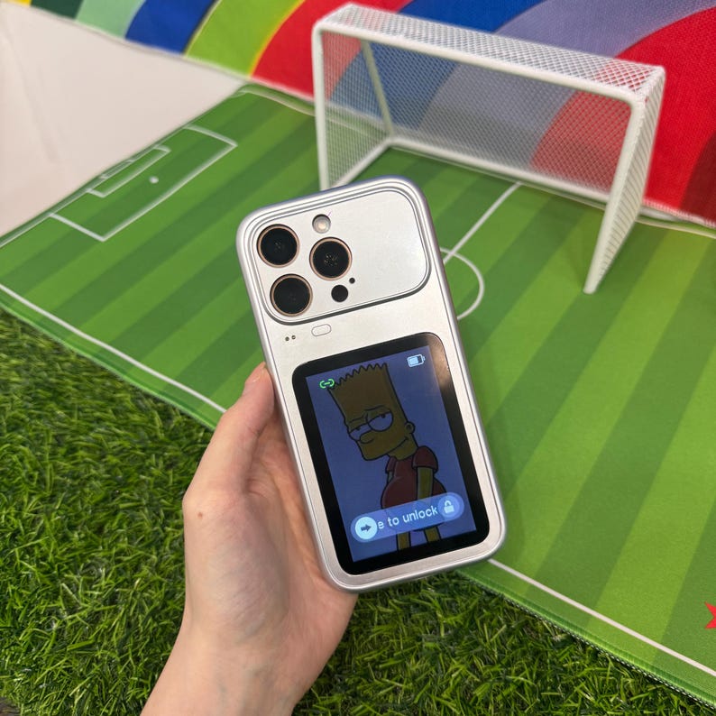 May include: Silver phone case with a Bart Simpson graphic on the screen. The phone case has a rectangular screen and a camera module with multiple lenses. The background shows a soccer field.